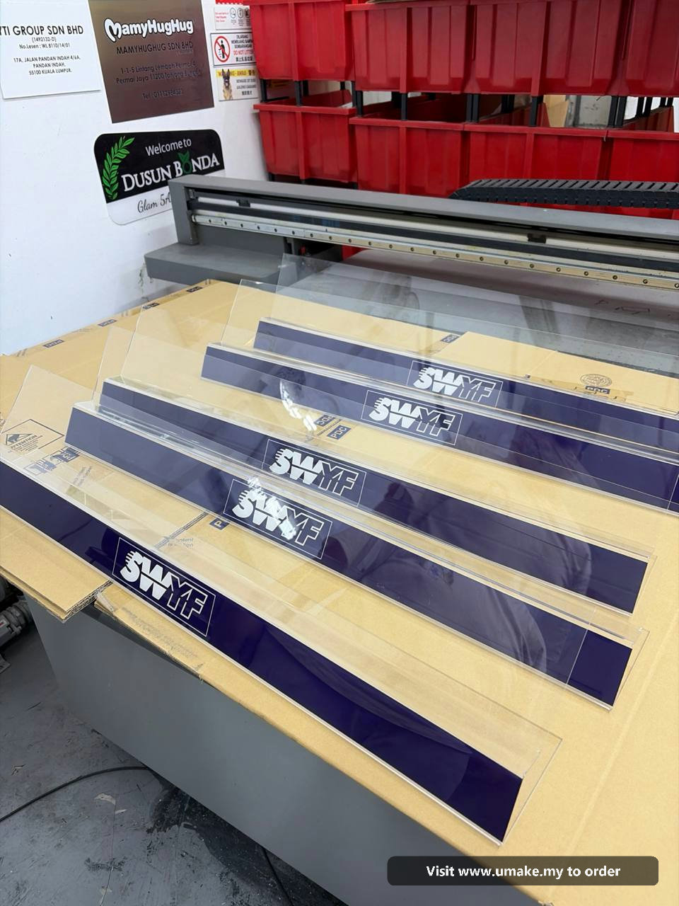 Custom Acrylic Display Stands by UMAKE Malaysia Several clear acrylic display stands with dark blue bases, featuring the white 'SWYF' logo, laid out on a cardboard surface in a manufacturing facilit