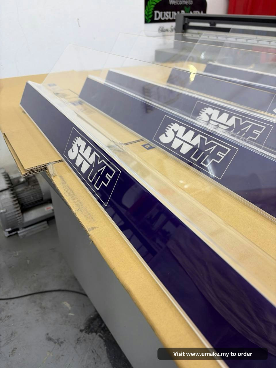 Custom Acrylic Display Stands by UMAKE Malaysia (2) Several clear acrylic display stands with dark blue bases, featuring the white 'SWYF' logo, laid out on a cardboard surface in a manufacturing facilit